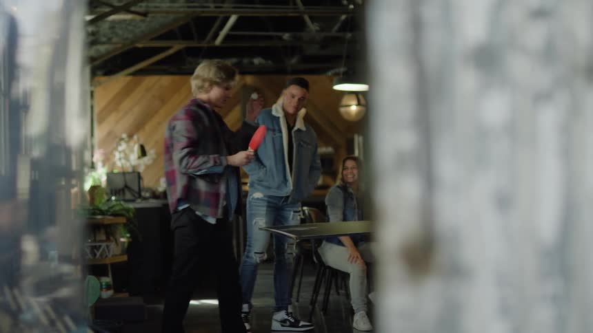 A small group of people are gathered around a table as two people play table tennis in a cafe