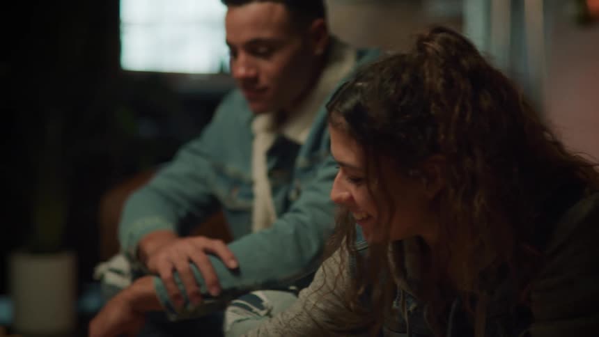 A teenage girl sitting next to a boy laughs as she picks up a card while playing a game
