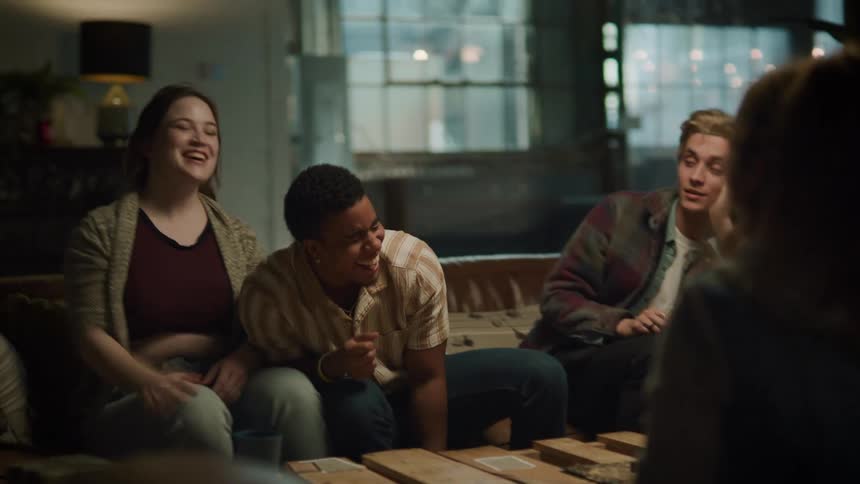Friends sitting on brown couch play cards and doing tricks in living room at home laughing