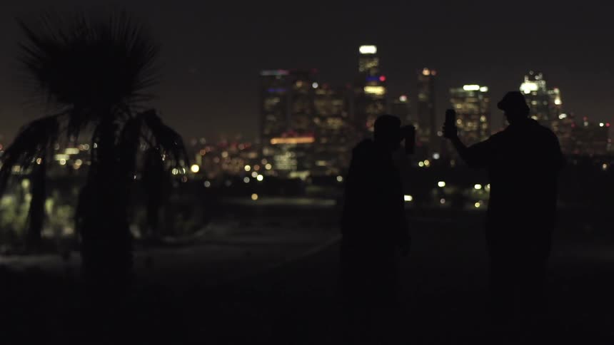 Two men toast each other with beer cans at night with a large city in the background