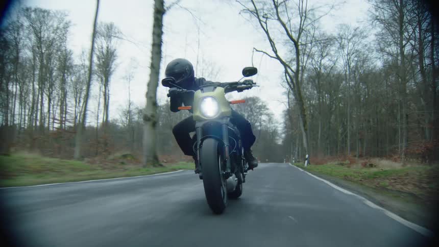 A cyclist wearing a black leather jacket and a helmet is riding a motorcycle on a rural road lined with trees