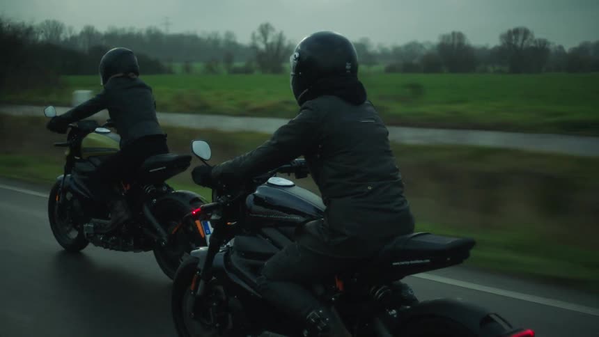 Two motorcycles traveling on a rural road past a stream and a wind turbine on a cloudy day