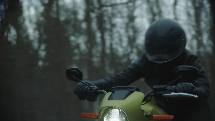 A cyclist wearing a black leather jacket and a helmet is riding a motorcycle on a rural road through the woods