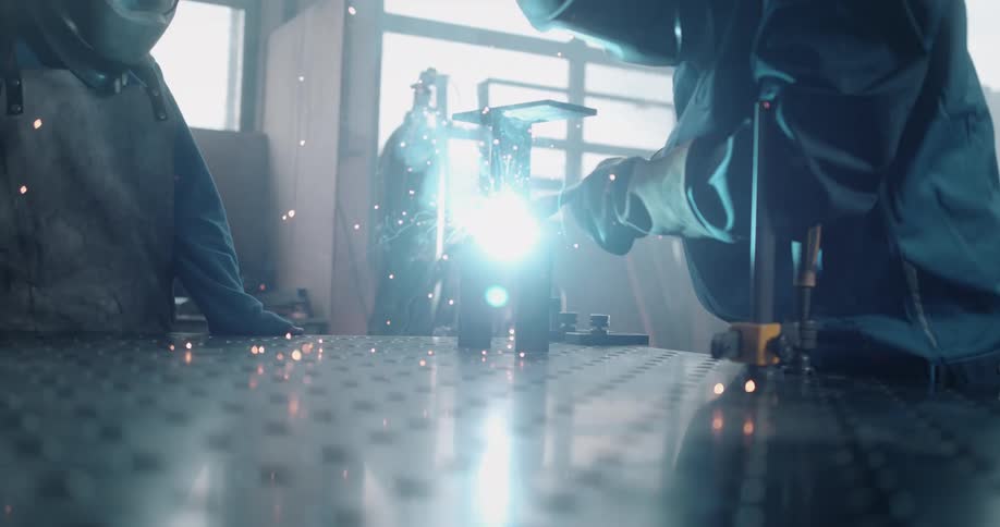 Two workers welding pieces of metal over a workbench at a factory