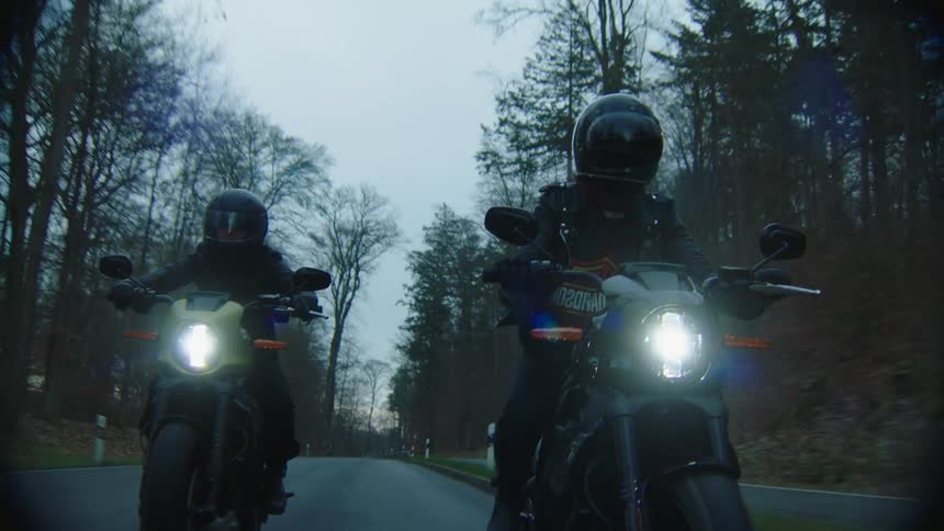 Two people ride motorcycles on the road past the forest