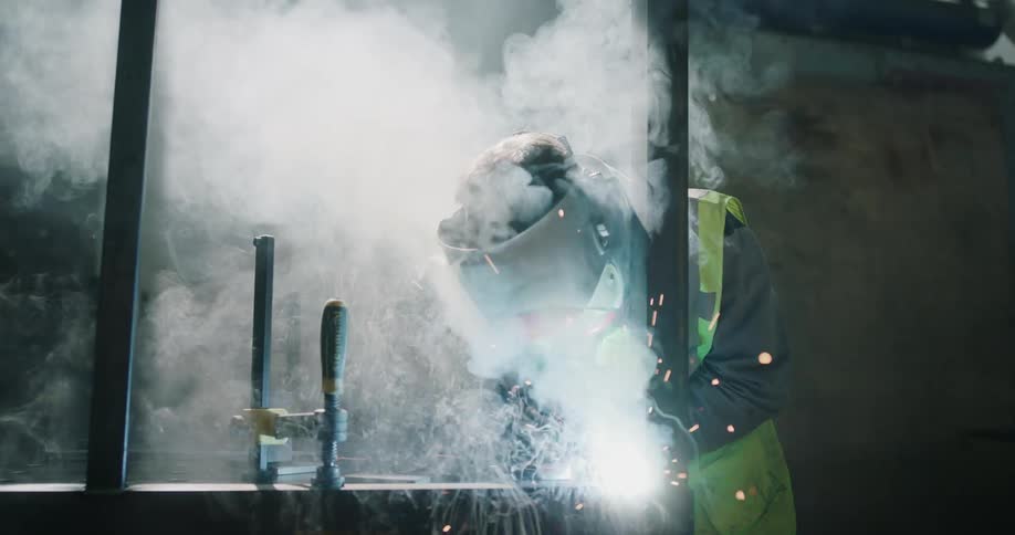 A welder is standing over a work area welding metal as sparks fly and smoke rises