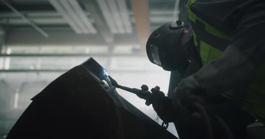 Welder wearing protective gearleans over at awkward angle to weld joint