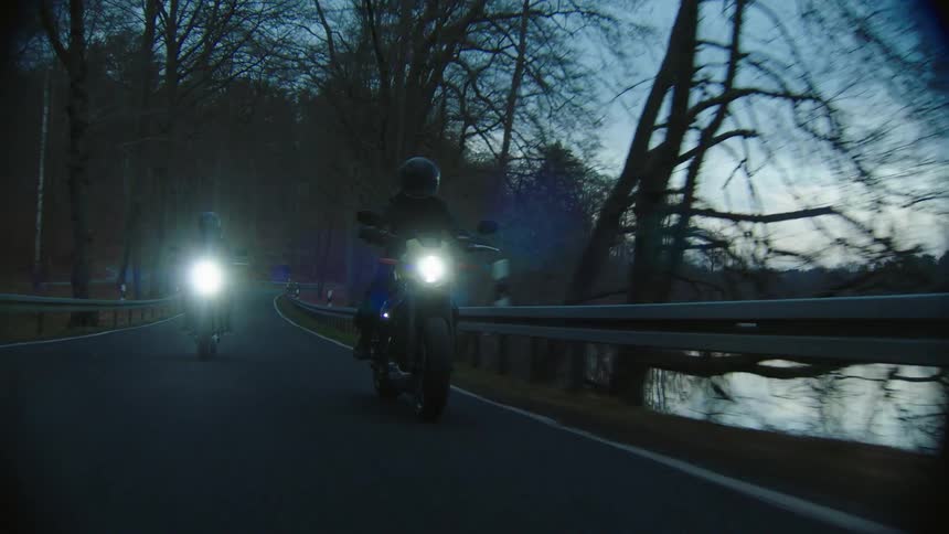 Two people ride motorcycles on a road near a river at evening