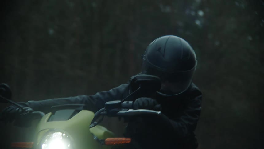 A cyclist wearing a black leather jacket and a helmet is riding a motorcycle on a rural road through the woods