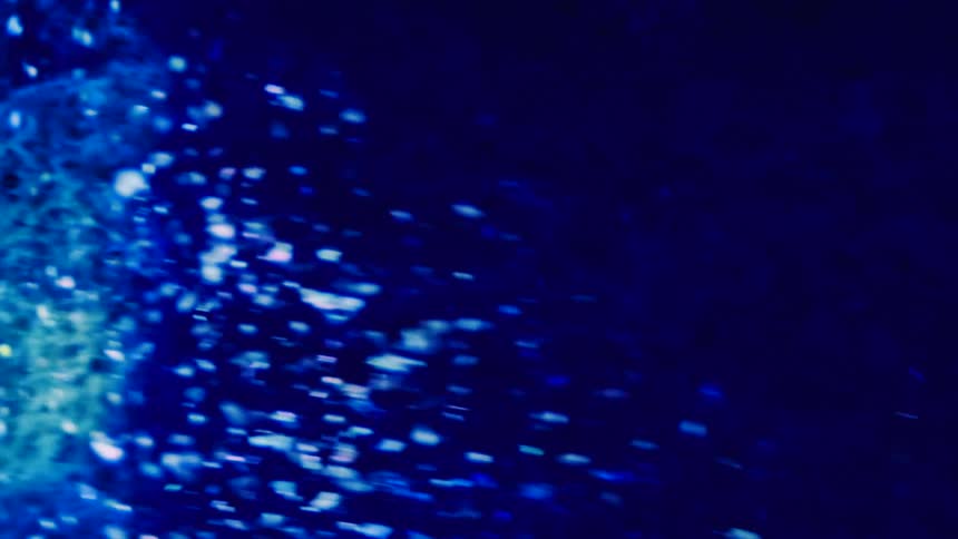 Green and white particles float in a yellow substance as an electric blue wave rushes over and light reflects on the bubbles inside