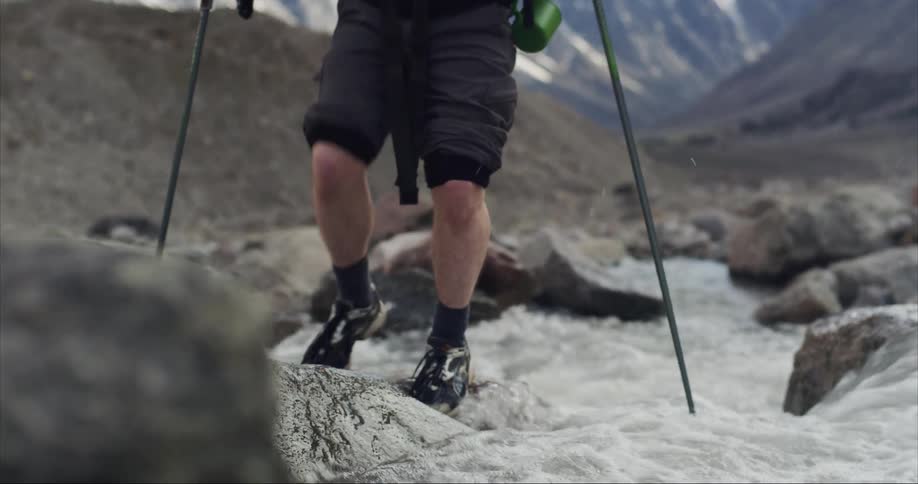 A man hiking with trekking poles makes his way across the rocks over a flowing stream and there are towering mountains in the distance