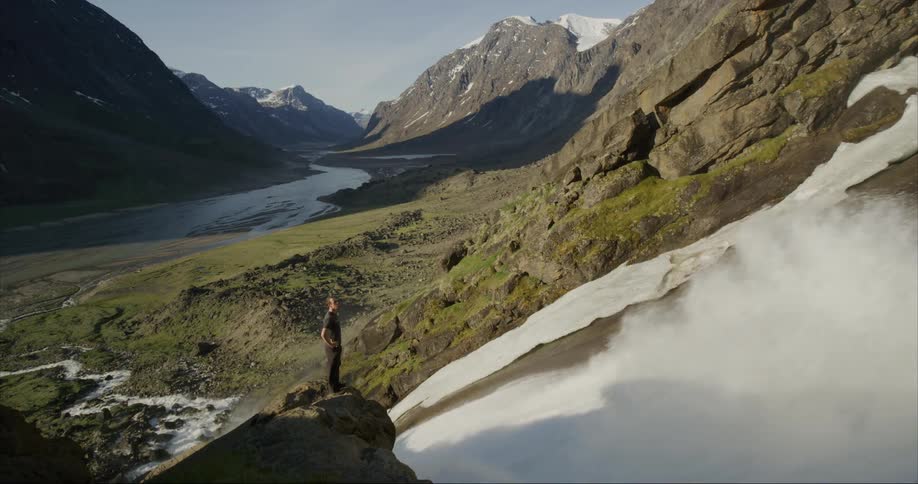 A man standing on a boulder overlooking a raging waterfall in a glacial river valley under the midnight sun