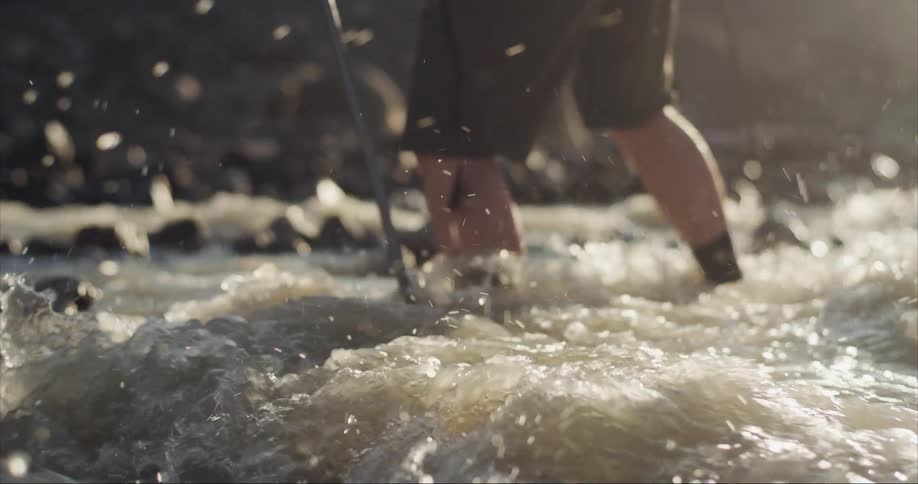 Man with brown shorts and black socks and shoes crosses a frigid mountain stream at sunrise