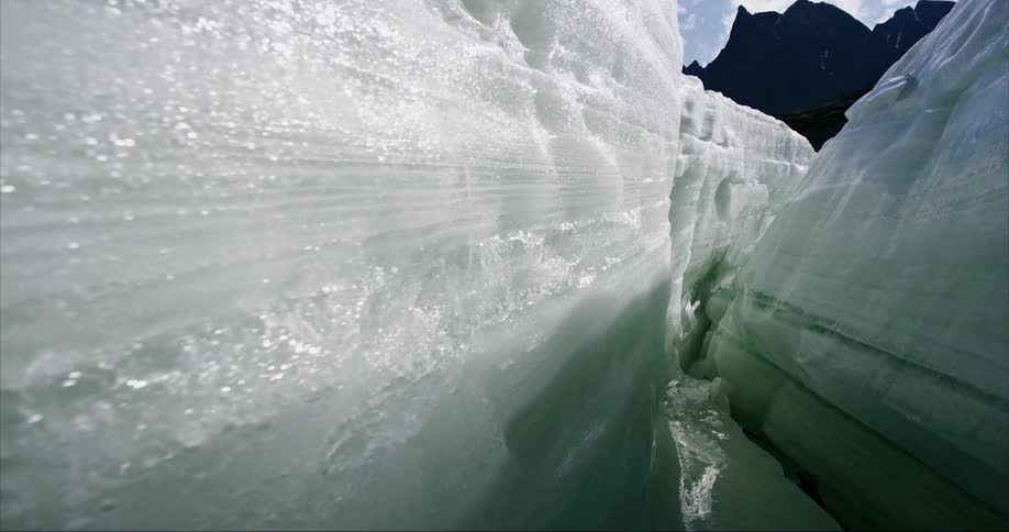 Sunlight shines on the ridges of a melting iceberg and there are mountains in the distance as the camera moves into the scene