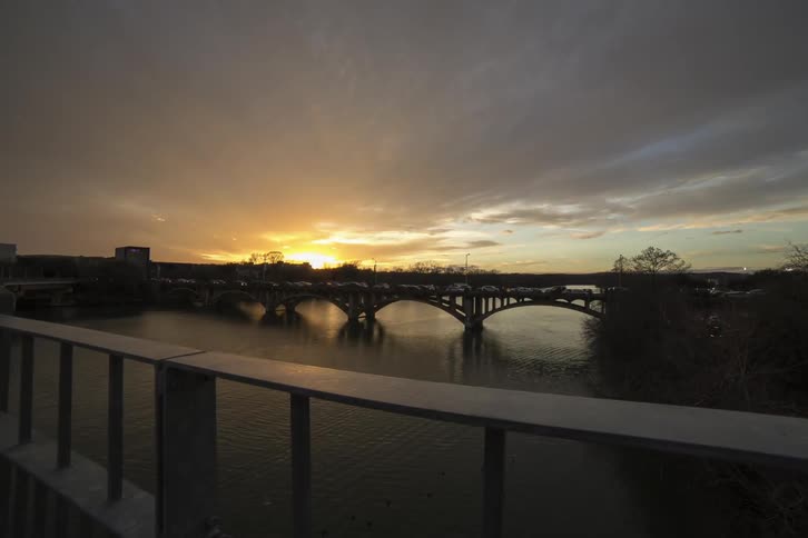 Traffic crossing a bridge that spans a river in an urban area as clouds stream overhead during sunrise