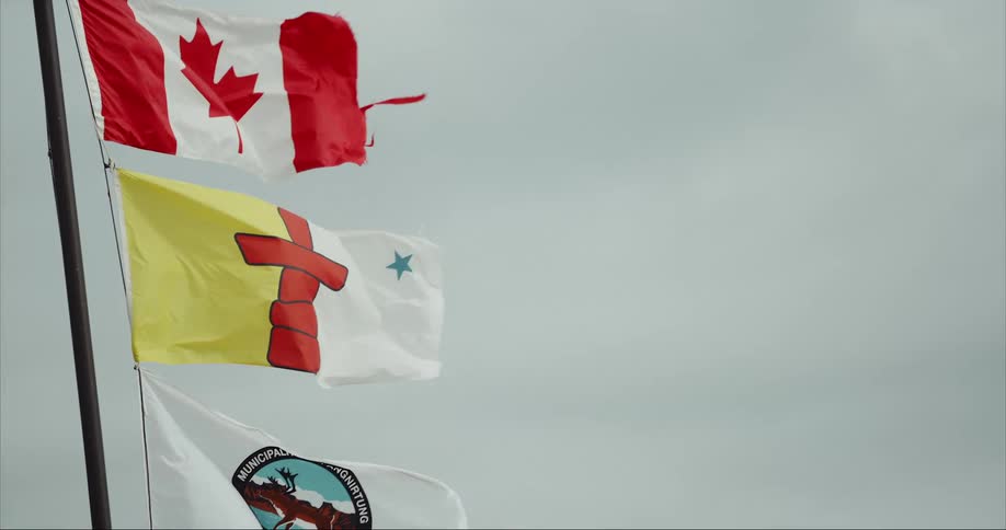 Three flags, Canadian, Nunavut and Municipality of Pangnirtung, waving in the wind with a car moving in the foreground