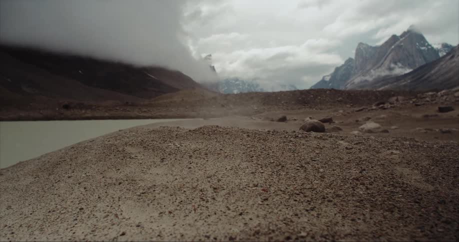 Dust flies near a terminal moraine lake as dark clouds move over the jagged glacier covered mountains