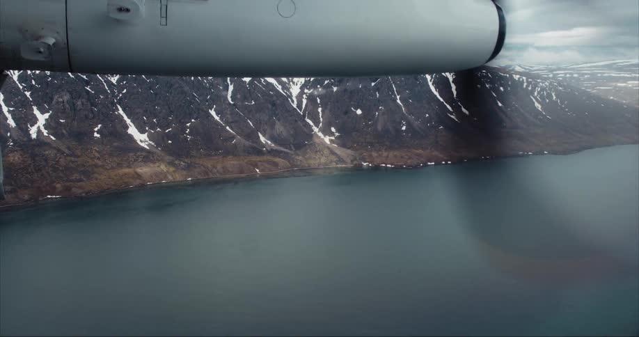 The propeller of a Dash 8 aircraft spins as it flies over water near glacier covered mountains