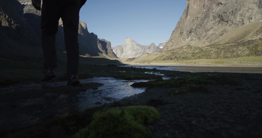 Man with pants and shoes crosses a stream in the glacier carved river valley with Mount Thor in the background