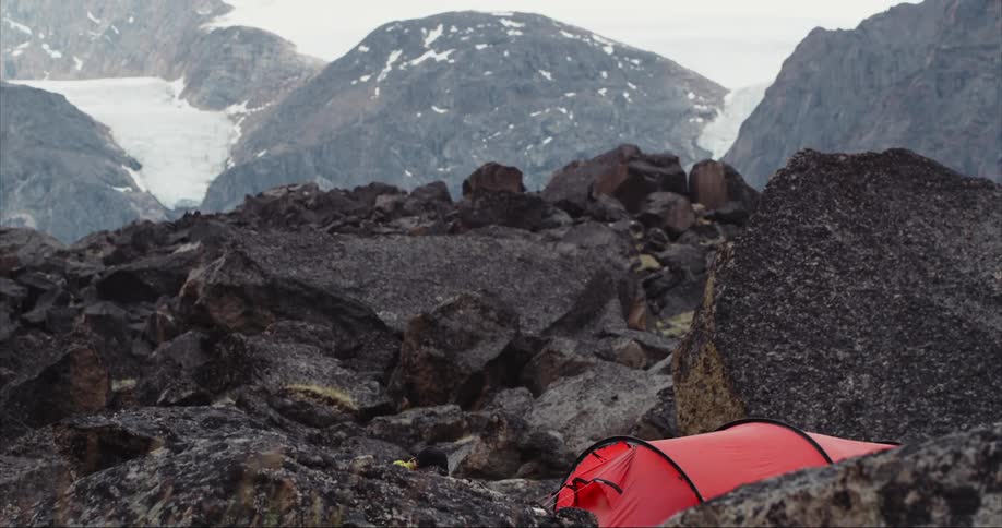 A tent is set up on a rocky mountain top near snow and glaciers in a rugged glacial valley