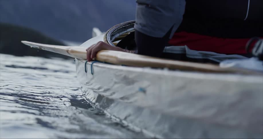 A person holds onto an paddle on the side of a traditional built sea kayak as it rocks on the waves and water drops fall gracefully into the Arctic Ocean