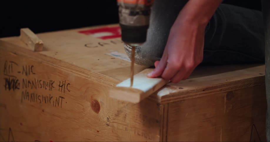 A person using an electric drill to make a hole in a piece of wood while holding it down with their leg