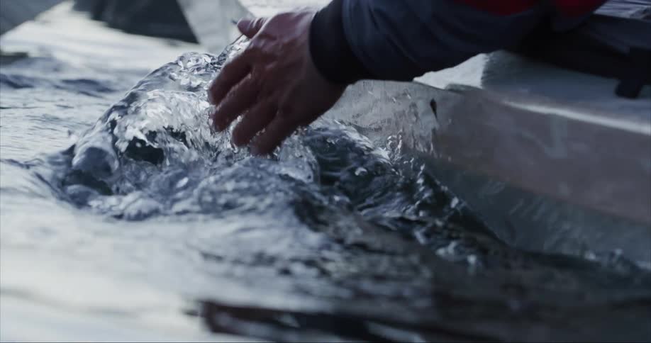 A man is leaning over in a sea kayaking and putting his hand in the water and splashing as the reflection from the light shines