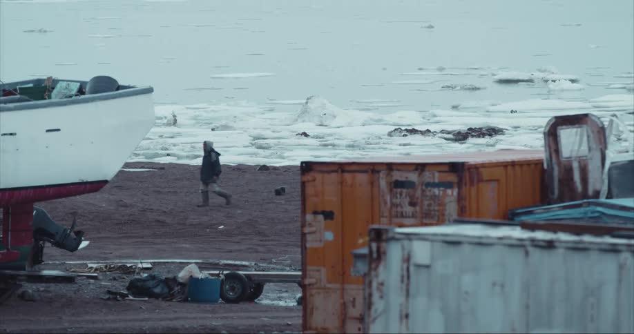 A person walking by shipping containers, dry land boat storage and sea ice on Baffin Island