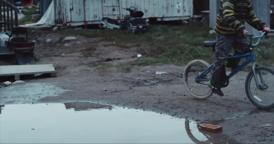 Young boy rides his bike casting a reflection on a puddle in front of his house on Baffin Island, Nunavut