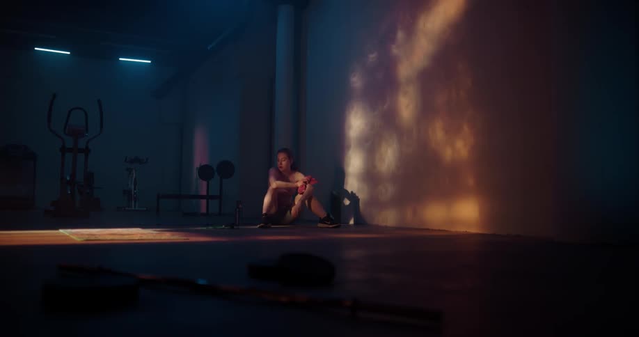 A woman sitting on the floor of a fitness room drinking from a water bottle