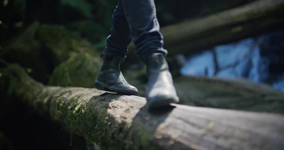 A person is walking across a fallen tree in the woods near a flowing stream