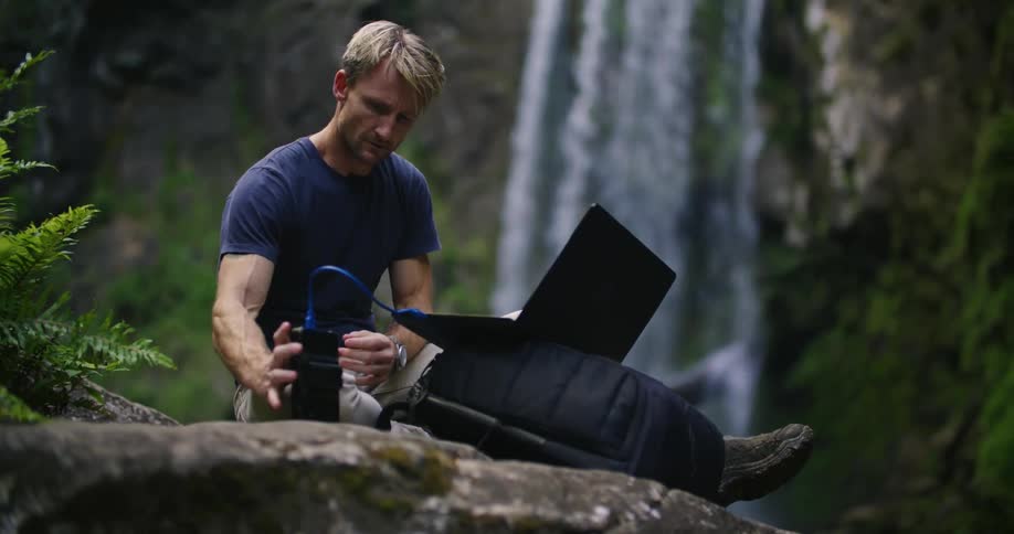 A man is sitting near a waterfall as he works on his computer and cellphone
