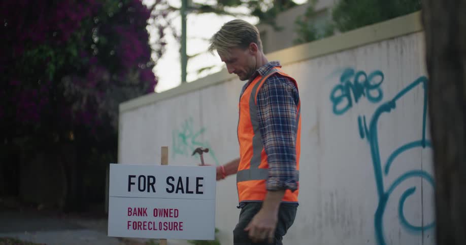 Worker hammers for sale sign for bank owned foreclosure into ground near wall with graffiti