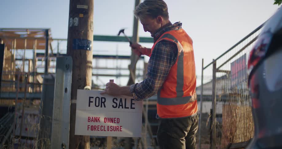 A man hammers a for sale sign outside a foreclosed property under construction
