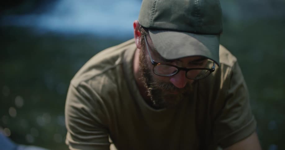 Man with green cap sunglasses beard and green t shirt takes photo of fungus near the river in the forest