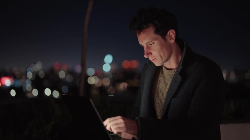Man working on a laptop on rooftop in front of city lights