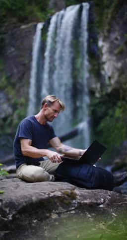 A man is typing on his laptop near a waterfall then reaches into a bag and pulls out a pen