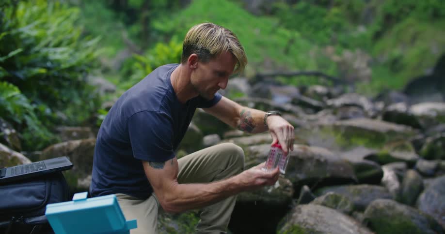 A man is kneeling next to water and fills a specimen bottle