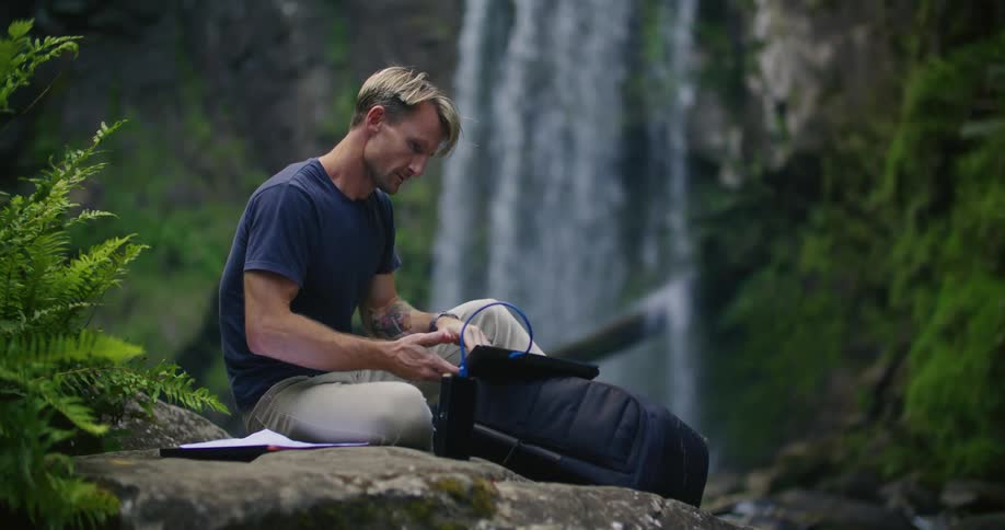 A man closes his laptop while sitting outside by a waterfall