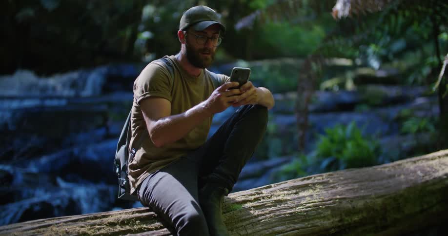 Man with cap glasses beard green shirt backpack and gray pants sits on tree trunk over stream using mobile phone in the forest