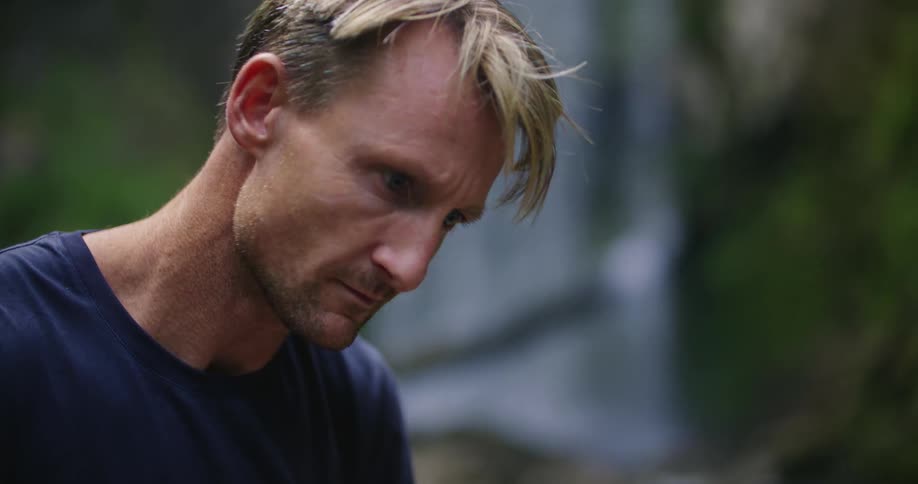 Man sitting on riverbank near waterfall typing on laptop stops briefly to rub his hand