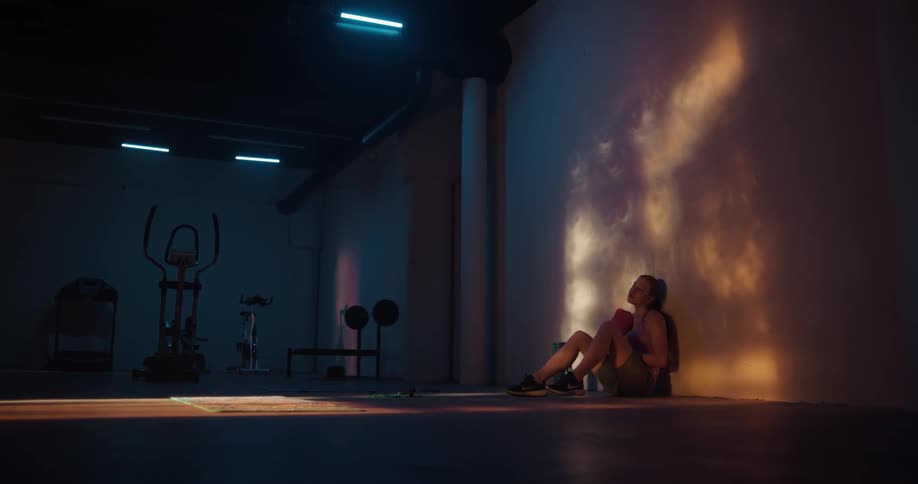 Woman wearing shorts and shirt sits against the wall drying her sweat in the gym