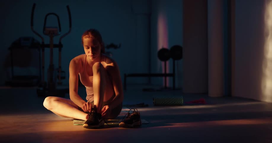 A woman is on a mat on the floor of a home gym putting on her shoes