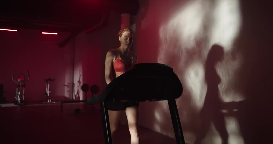 Woman walking on treadmill in dimly lit gym casts shadow on wall