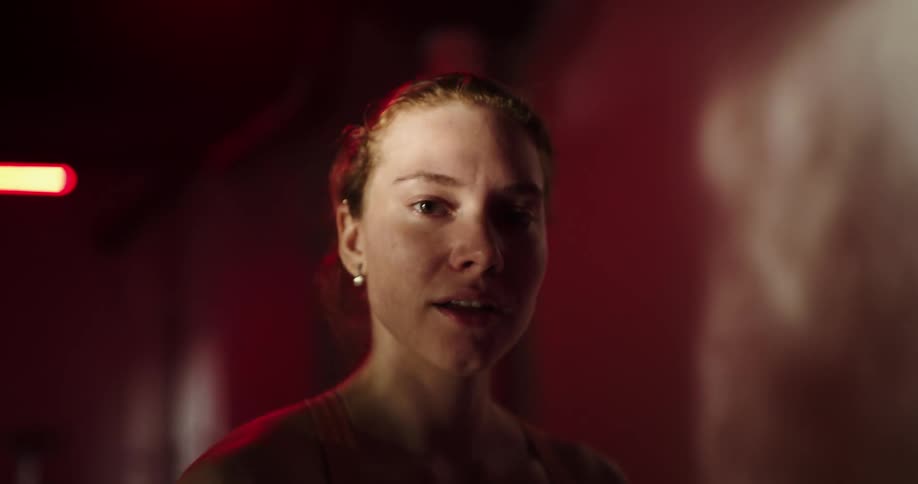 Woman with ponytail orange sports bra and black shorts exercises on treadmill and smiles at camera