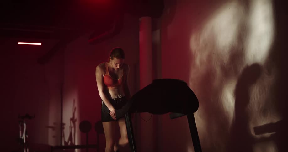 Woman with ponytail orange sports bra and black shorts hurts her knee while running on treadmill in a gym