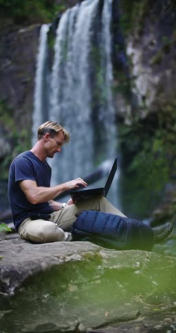 A man sitting near a waterfall clicks his pen then types on a laptop