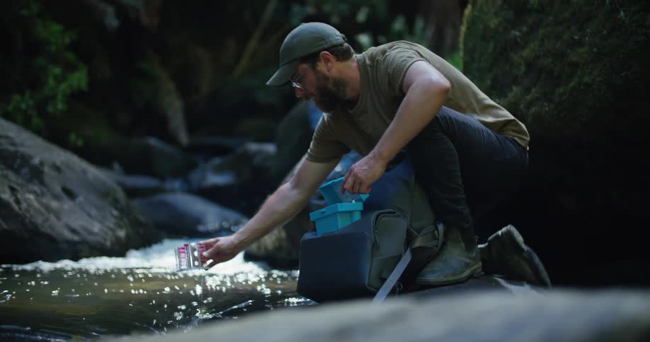 Man crouches near stream collecting water sample