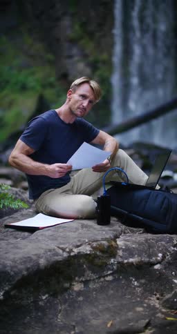 A man reads papers while sitting with his laptop near a waterfall