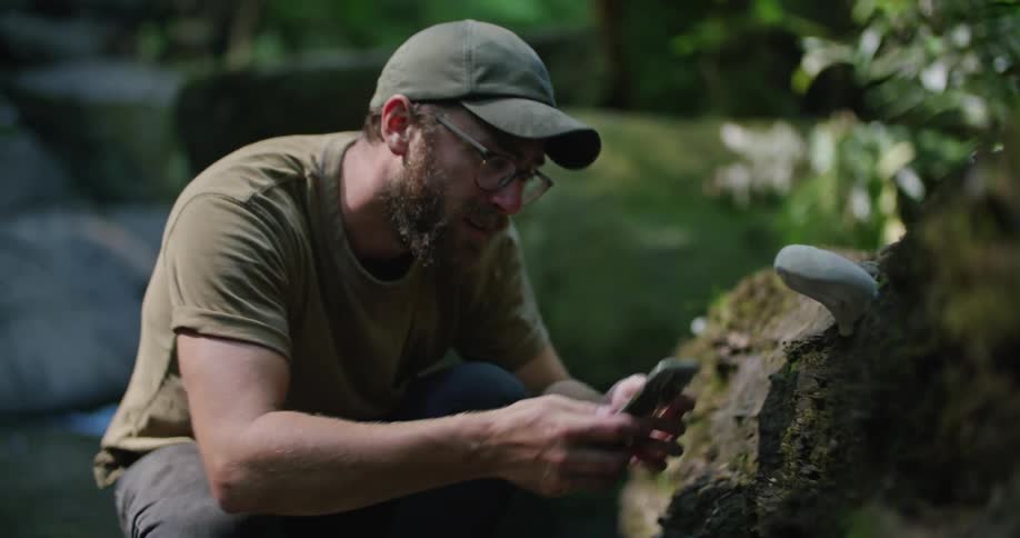 A man squats down next to a rock formation and takes a picture with his cell phone